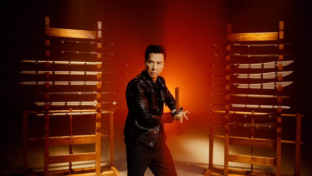 Donnie Yen standing between two racks of traditional Chinese weapons in an official Phantom Blade Zero promo shoot, wearing a black leather jacket and gripping a weapon with both hands